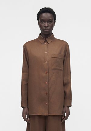 Czarna kobieta stojąca, ubrana w brązową koszulę z długim rękawem i kieszenią na piersi, na jednolitym jasnym tle.