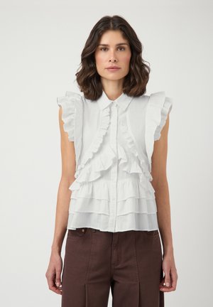 Kobieta z ciemnymi włosami do ramion, ubrana w białą, marszczoną, bezrękawnikową bluzkę i ciemnobrązowe spodnie, stojąca na jednolitym tle.