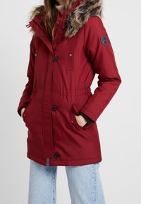 Röd parka-jacka med huva i fuskpäls, med en insvängd midja, knappstängningar och sidofickor. Tillverkad av texturerat tyg.