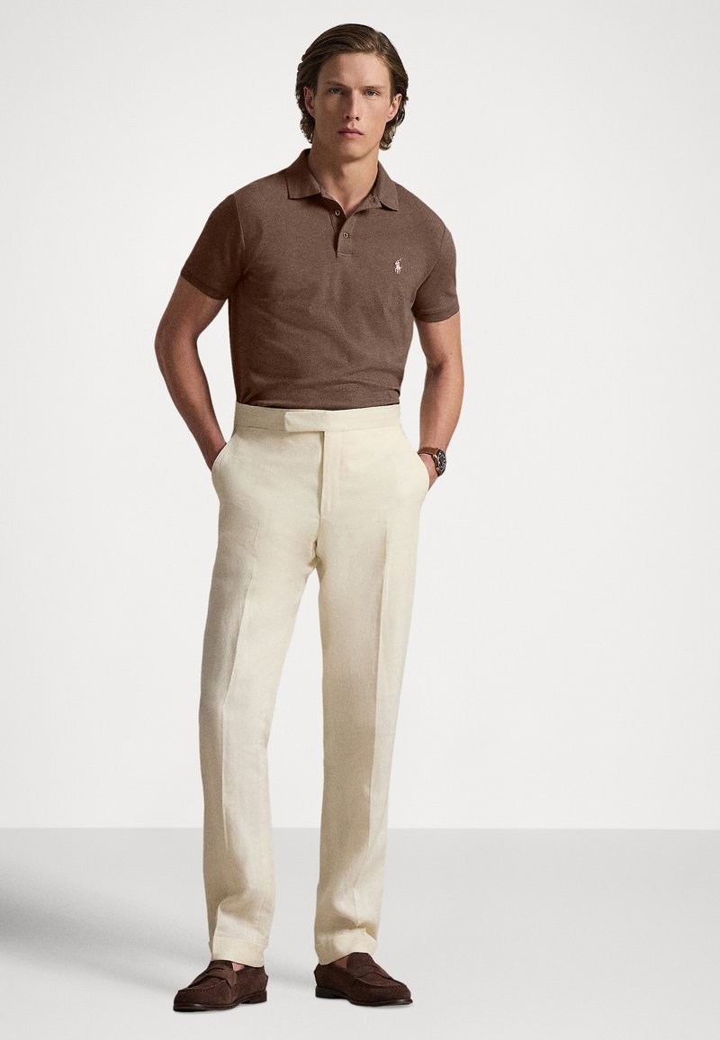 Braunes Polohemd mit kurzen Ärmeln, cremefarbene maßgeschneiderte Hose und braune Wildleder-Loafer. Glatte Textur; dezente Markenkennzeichnung auf der Brust.