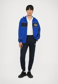 Plava zip-up dukserica s crnim naglascima i logotipom, uparena s tamnoplaviim trenirkama i crnim tenisicama. Glatka tkanina i ležeran kroj.