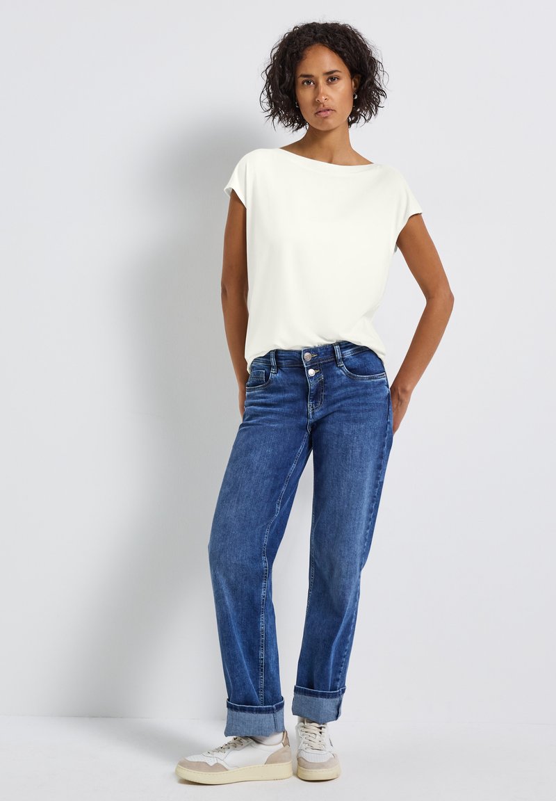 Vit kortärmad topp i kombination med blå denim jeans. Sneakers med beige och vita detaljer. Slät tyg med en avslappnad passform.