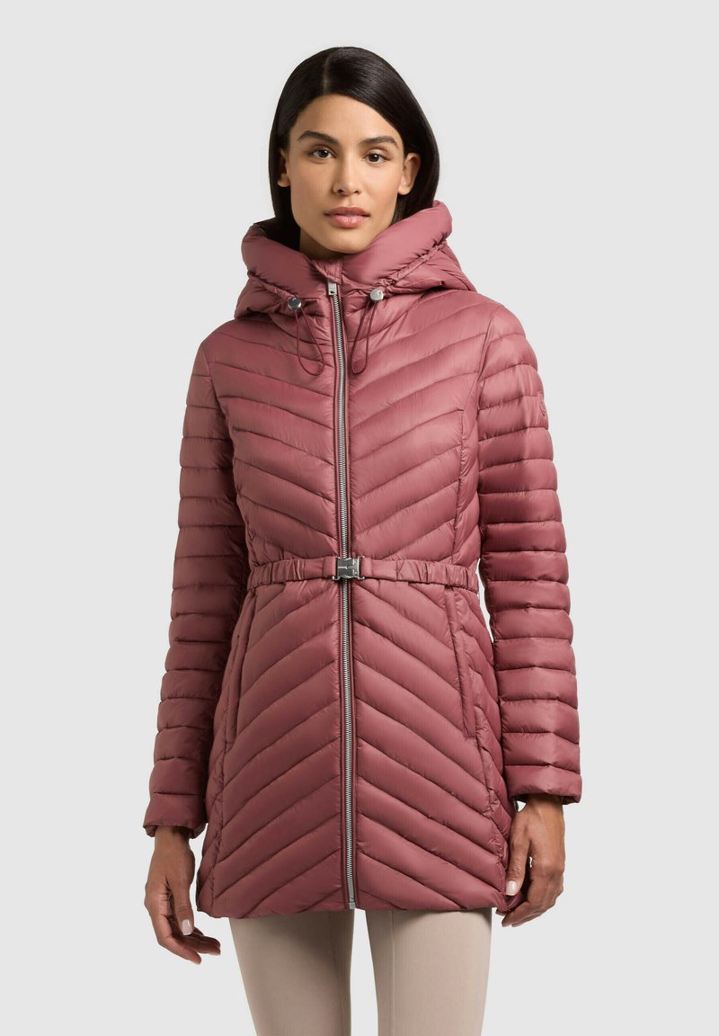 Naine kannab lavendlivärvi tikitud sulejopet kapuutsiga, vöökohaga ja esikinnitusega, seistes helehalli ühevärvilise tausta ees.