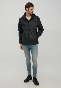 Giacca nera impermeabile con zip frontale, cappuccio regolabile e polsini elasticizzati. Indossata con jeans azzurri chiari e stivali neri con lacci.