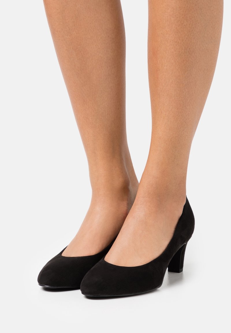 Deux jambes portant des escarpins mi-talons en daim noir, vus de la moitié du mollet vers le bas sur un fond uni clair.