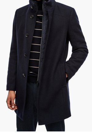 Homme portant un manteau bleu marine foncé avec boutons et fermeture éclair, une main dans la poche, par-dessus un pull rayé noir et blanc, sur fond blanc.