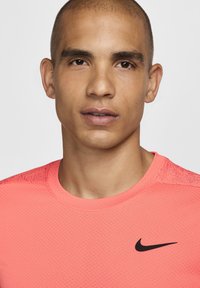 Nike trumpa rankovėmis marškinėliai ryškiai rožinės spalvos audinio su raštuotais paviršiais, apvaliu kaklu ir juodu logotipu kairėje krūtinės pusėje.