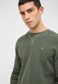 Õlivihreast tekstuuriga ümarkaelusega kampsun, mille rinnal on väike tikitud logo, tugevdatud kaelusega ja ristpistes detailidega.