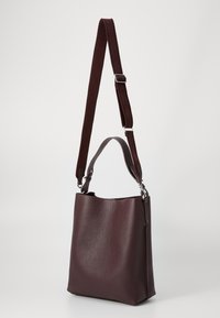 Sac en cuir bordeaux avec une finition texturée, une courte anse supérieure et une longue sangle réglable ornée de détails de logo. Forme globale rectangulaire.