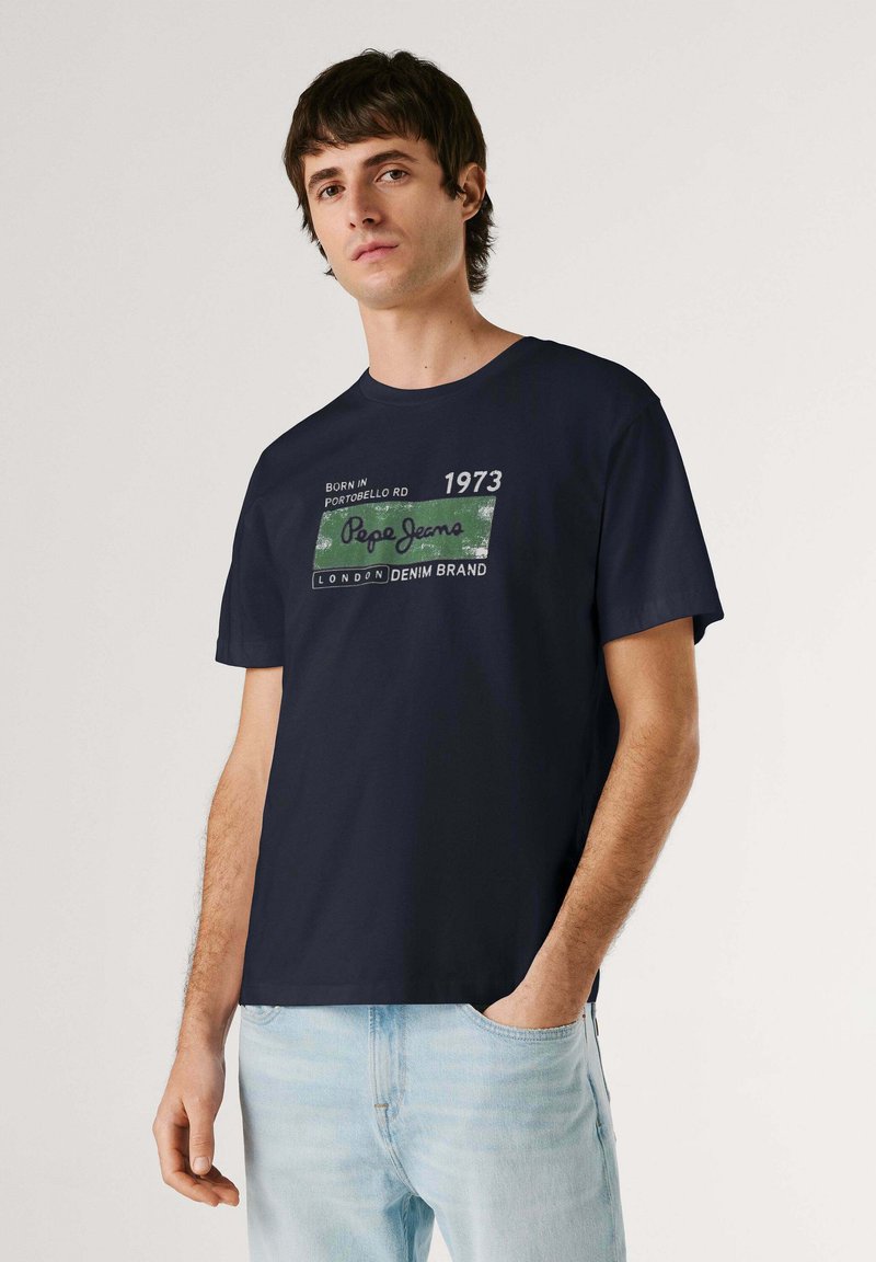 Jeune homme portant un t-shirt bleu marine foncé avec un motif texte vert et blanc, jean bleu clair, debout avec une main dans la poche devant un fond uni.