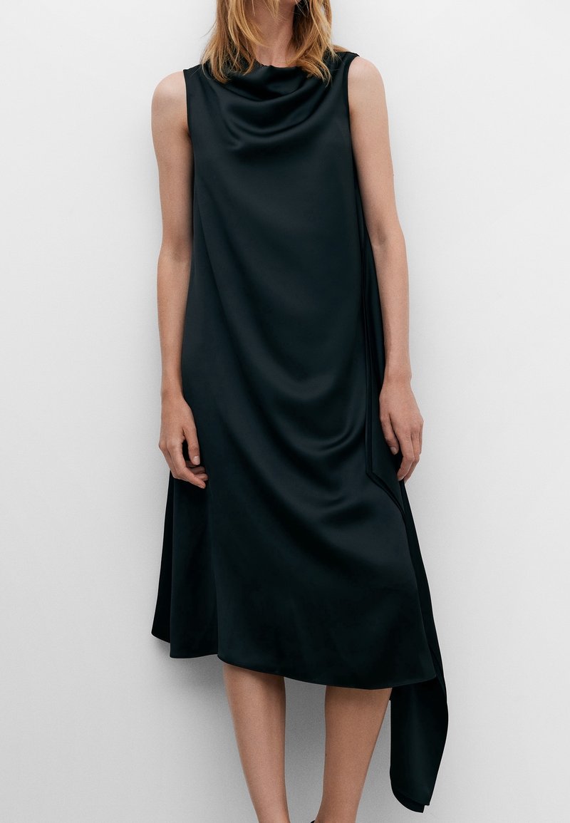 Robe sans manches noire en tissu lisse semblable à du satin, présentant une encolure à godets et un ourlet asymétrique avec un côté drapé.