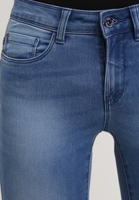 Modré džíny z denimu s hladkou texturou, středním pasem, přední kapsou a mírným blednutím směrem k kolenům. Obsahují zapínání na knoflík a poutka na opasek.