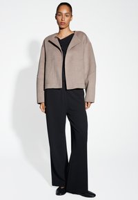 Giacca cropped in misto lana beige con maniche oversize, chiusura a singolo bottone e due tasche frontali, abbinata a pantaloni neri a gamba larga.