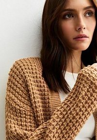 Cardigan lavorato a maglia in marrone chiaro, con un motivo testurizzato, apertura frontale e maniche lunghe, indossato sopra una maglietta bianca.