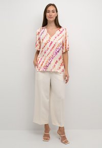 Blusa leggera in crema con strisce diagonali rosa e arancioni; scollatura a V; maniche ampie. Abbinata a pantaloni larghi in crema e tacchi con cinturini.