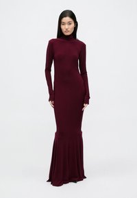 Burgundia värvi pikkade varrukatega kleit kõrge kaelusega ja lehvivate äärega, valmistatud siledast, venivast materjalist, mis järgib keha kujusid.