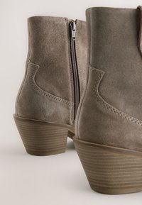 Brune ruskinds-ankelstøvler med en sidelukning med lynlås, stablet hæl og syninger som detaljer. Hælen har vandrette linjer for tekstur.