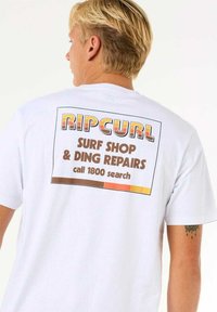 T-shirt bianco in cotone con una grafica sul retro che presenta la scritta "RIPCURL" in testo multicolore. Il design include dettagli come "SURF SHOP & DING REPAIRS."