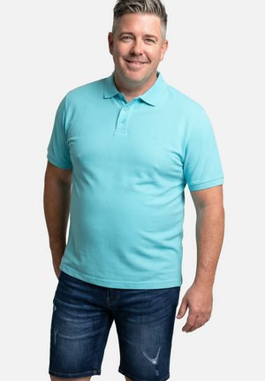 Mężczyzna uśmiechający się, ubrany w jasnoniebieską koszulkę polo i ciemnoniebieskie szorty dżinsowe, stoi z rękami swobodnie opuszczonymi wzdłuż ciała na białym tle.