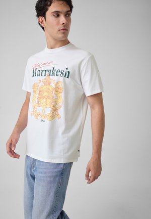 Jeune homme portant un t-shirt blanc avec l’inscription ‘Marrakesh’ et un motif blason, associé à un jean bleu clair, debout devant un fond gris uni.