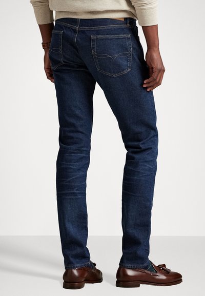 Dark blue denim jeans with a slim fit, featuring double back pockets, visible stitching details, and a straight leg design.