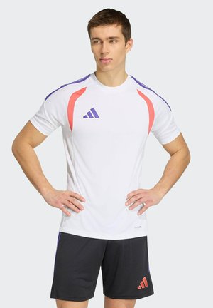 Młody mężczyzna stoi z rękami na biodrach, ubrany w biały sportowy T-shirt z fioletowymi i czerwonymi akcentami oraz czarne spodenki z czerwonym logo.