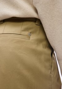 Μπεζ πουλόβερ μπροστά στη μέση σε παντελόνι καμπάνα χρώματος χακί, εμφανίζοντας την πίσω τσέπη με μικρό ασημένιο λογότυπο της μάρκας στη δεξιά πλευρά.