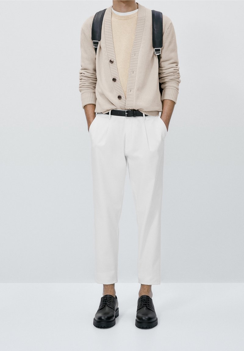 Cardigan beige en maille avec des boutons, pull crème en dessous, pantalon plissé blanc et chaussures noires à lacets, porté avec un sac à dos noir.