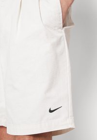 Detailní záběr osoby v světlobéžových šortkách s černým logem Nike swoosh blízko lemu a rukama v kapsách.