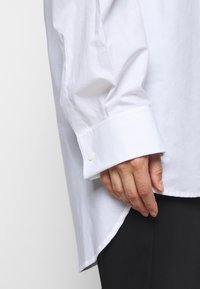 Vit skjorta med lång ärm, som har en manschett med knappstängning och en lätt böjd nederkant. Slät tygstruktur.