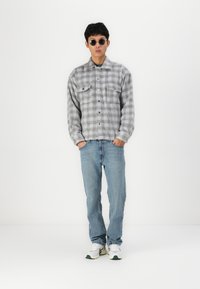 Lichtgrijs geruit overhemd met twee voorzakken, gecombineerd met lichtblauwe jeans en witte sneakers. Accessoires zijn ronde zonnebrillen.