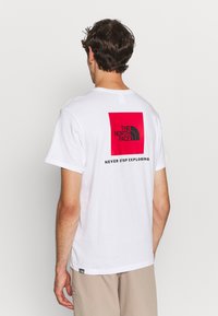 Camiseta blanca de algodón con un gran diseño de cuadrado rojo en la parte posterior que dice "THE NORTH FACE" y "NEVER STOP EXPLORING" en texto negro.