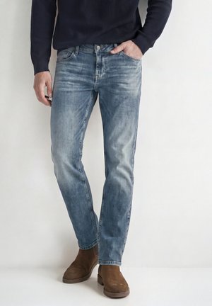 Homme portant un jean bleu délavé, des bottes en cuir marron et une chemise sombre à manches longues, debout avec une main dans la poche sur un fond blanc.