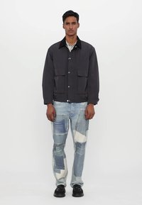 Férfi sötétszürke dzseki négy elülső zsebbel, világoskék foltos farmernadrággal párosítva, amely különböző árnyalatú denimből készült, és fekete cipővel.