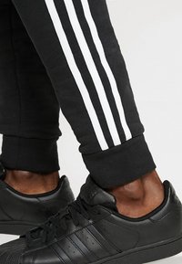 Svarta Adidas-sneakers bärs med svarta byxor med tre vita ränder längs sidan, visar vrister och fötter på en neutral bakgrund.
