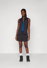 Colete preto sem mangas com fecho de correr, combinado com uma blusa azul e uma saia preta. Tecido texturizado e detalhes de bolso em destaque. Calçado preto.