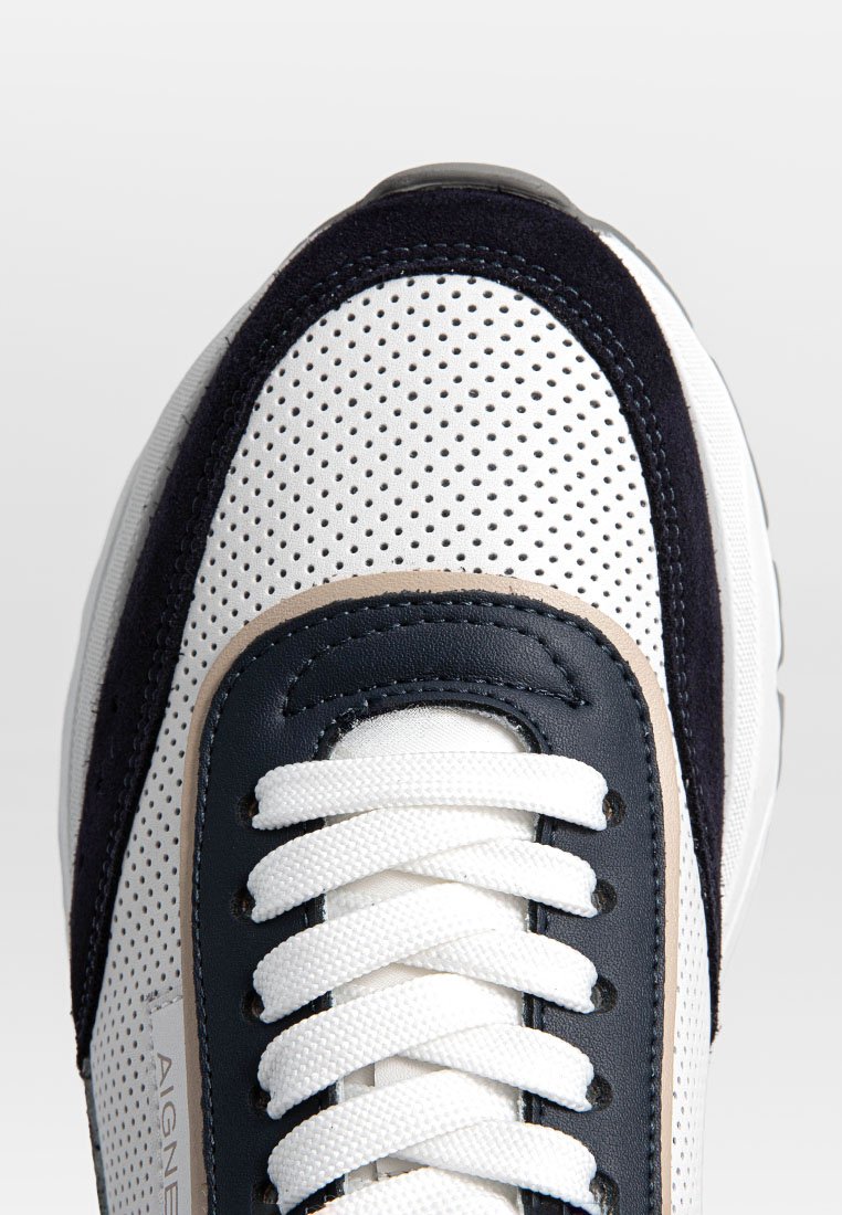 Weiße Sport-Schuhspitze mit perforiertem Obermaterial, marineblauen und beigen Lederakzenten sowie weißen Schnürsenkeln vor einem schlichten hellgrauen Hintergrund.