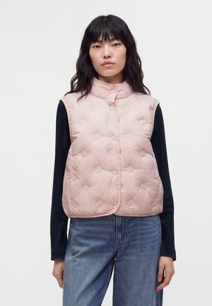 Femme aux cheveux noirs portant un gilet matelassé rose clair sur un haut noir à manches longues et un jean bleu sur un fond blanc uni.