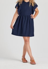 Granatowa sukienka z oczkami, z falbankami na rękawach i marszczonym pasem. Połączona z brązowymi sandałami na ramiączkach. Gładki materiał z wycięciami.