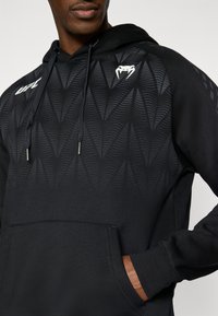 Μαύρο φούτερ με κουκούλα με υφή σε σκούρες αποχρώσεις, κλεί σιμο με κορδόνι και τσέπη καγκουρό. Λεπτομέρειες λογότυπου είναι ορατές στο στήθος.