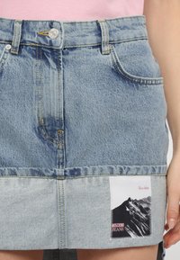 Fakó kék tónusú farmer mini szoknya, öt zsebes kialakítással, és hegyeket valamint a márka logóját ábrázoló grafikus folttal az alján.