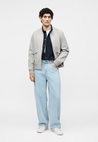 Ung mand stående iført en lysegrå bomberjakke, sort skjorte med knapper, lyseblå vidbenede jeans og hvide sneakers.