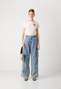 Beige t-shirt with embroidered logo, paired with wide-legged blue denim cargo pants with pocket details. Silver handbag and metallic shoes.