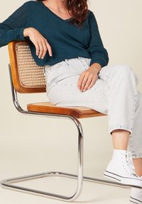 Femme assise sur une chaise en bois et cannage avec des pieds chromés, portant un haut à manches longues bleu sarcelle, un jean bleu clair et des baskets blanches.