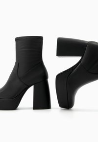 Černé kotníkové boty s hladkým koženým povrchem, vysokým blokovým podpatkem a bezproblémovým designem, zvýrazněné mírně vyvýšenou podrážkou.
