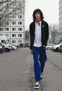 Black zip-up jacket with white stripes, white t-shirt, red bandana, blue wide-leg jeans, and black high-top sneakers. Urban street setting.