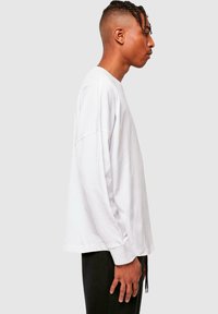 Langærmet hvid bomulds t-shirt med en afslappet pasform, nedfalden skulder og en lige kant, båret med mørke bukser, set fra siden.
