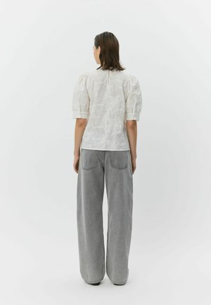 Vrouw met kort donker haar, gekleed in een witte blouse met gestructureerde pofmouwen en losse grijze jeans, staand voor een effen witte achtergrond.
