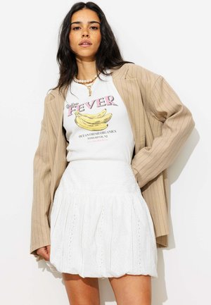 Femme aux longs cheveux foncés portant un blazer beige à rayures fines, un t-shirt blanc avec un motif de bananes et une jupe blanche brodée, debout devant un fond blanc.