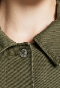 Olivově zelená bavlněná bunda se strukturovaným límcem a viditelnou texturovanou tkaninou. Na výstřihu má kulatý černý knoflík.